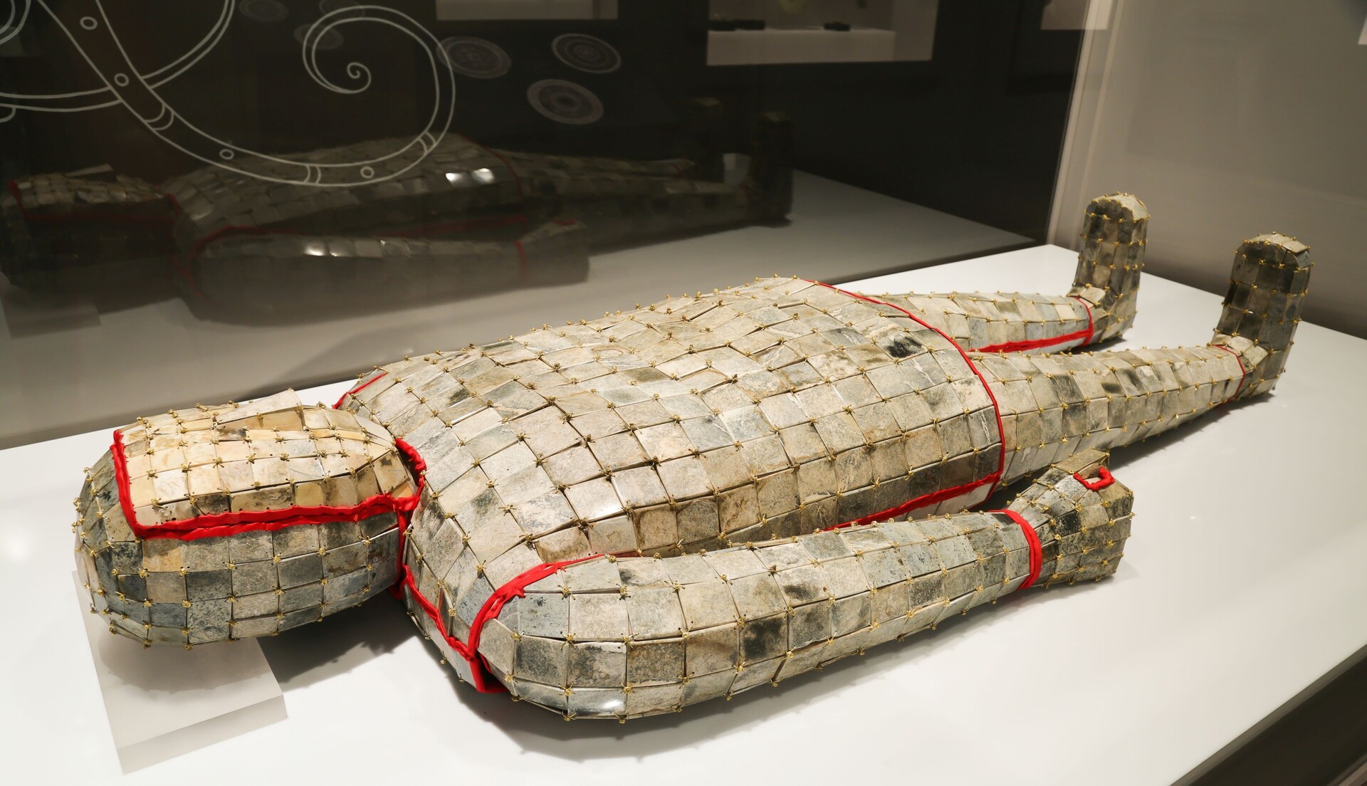 A complete jade burial suit lying in a display case, formed from rectangular jade plaques joined with gold wire and edged with red textile.